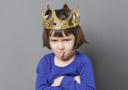 spoiled kid concept - cheeky preschool child with golden crown on head folding arms and sticking out tongue for disrespectful mollycoddled little king or queen metaphor, long grey banner