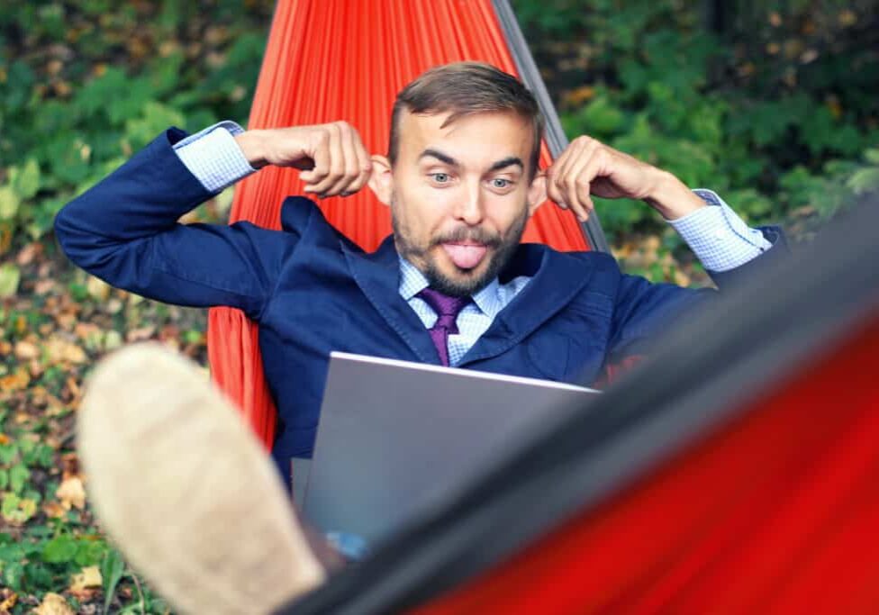 Businessman with laptop relaxs in a hammock on nature. He talking on video chat and doing funny faces with his tongue hanging out and splayed ears. Freelance or telework, on-line chatting concept.